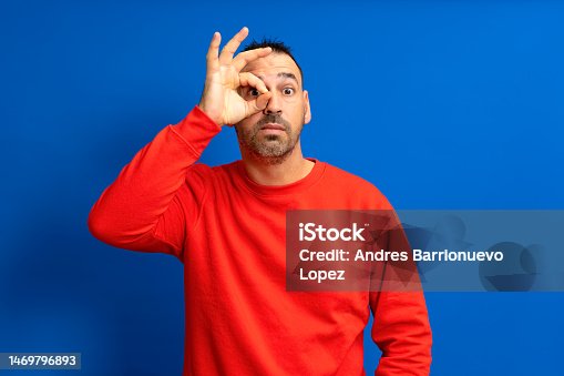 istock Bearded hispanic man in his 40s wearing a red jumper making the gesture of looking through a telescope by one hand isolated on blue studio background. 1469796893