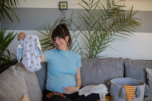 Young happy pregnant woman is sitting on the couch in her home. She is smiling and arranging clothes for her baby.