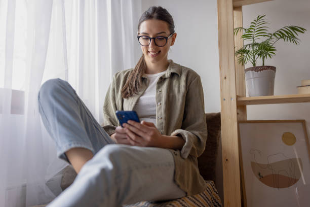 young vestida casualmente usando el teléfono mientras se relaja en su apartamento - young women sitting simple living eastern europe fotografías e imágenes de stock