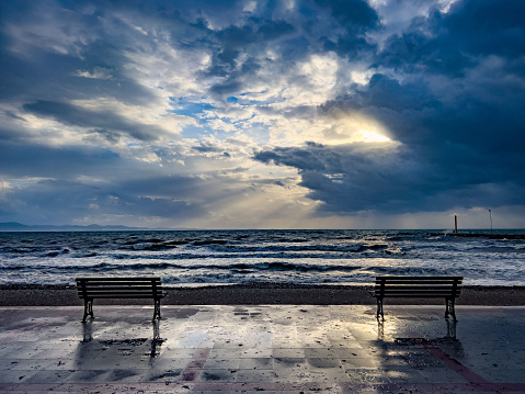 Powerful dramatic storm over ocean beach with golden light. View from the shore on sea, sky, setting sun and clouds. Silhouette of bench in front of water. Beautiful nature scenery background with copy space. Incredible sunset beneath dark dramatic clouds.