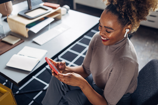 A from above view of a smiling African-American entrepreneur listening to music using her wireless earbuds and her smartphone. She is sitting at the desk in her office.