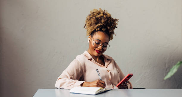 istock A Happy Beautiful Businesswoman With Blonde Curly Hair Taking Some Notes While Using Her Earbuds And Mobile Phone 1469817370