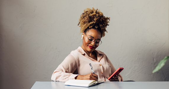 A smiling African-American entrepreneur with glasses writing something in her notebook while listening to some business podcast on her smartphone. She is sitting at the desk in her office. (copy space)