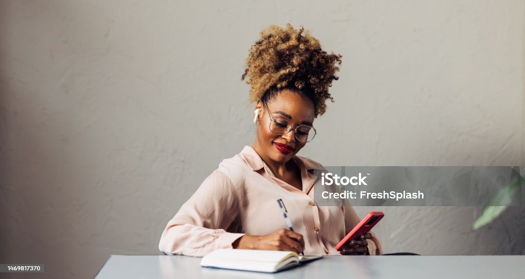 A Happy Beautiful Businesswoman With Blonde Curly Hair Taking Some Notes While Using Her Earbuds And Mobile Phone A smiling African-American entrepreneur with glasses writing something in her notebook while listening to some business podcast on her smartphone. She is sitting at the desk in her office. (copy space) Women Stock Photo