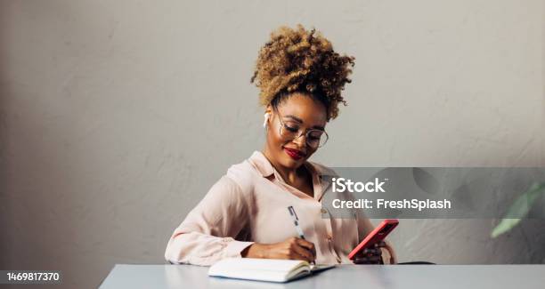 A Happy Beautiful Businesswoman With Blonde Curly Hair Taking Some Notes While Using Her Earbuds And Mobile Phone Stock Photo - Download Image Now