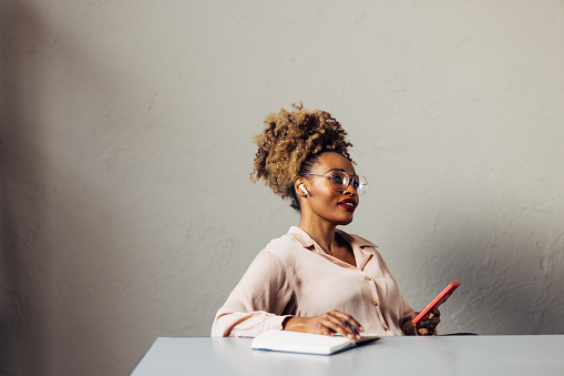 A smiling African-American entrepreneur with glasses writing contemplating while listening to some business podcast on her smartphone. She is sitting at the desk in her office. (copy space)