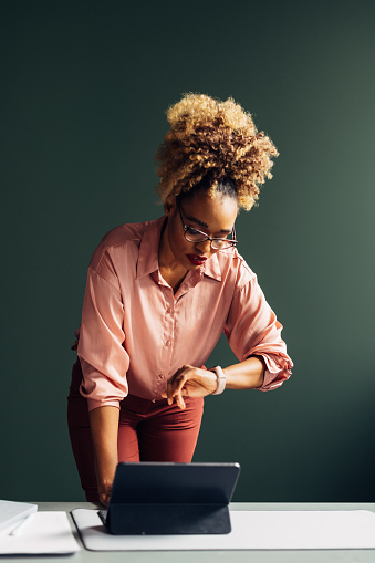 An angry African-American entrepreneur with glasses looking at her smartwatch while waiting for somebody. She is standing at the desk in her office. (copy space)