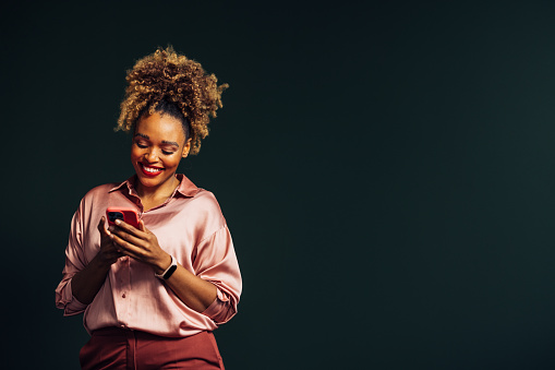A smiling elegant African-American entrepreneur chatting on her smartphone while standing against grey background. (studio shot, copy space)