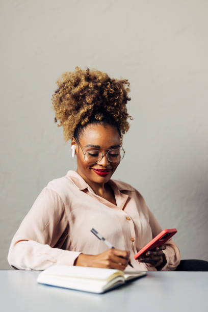 A Happy Beautiful Businesswoman With Blonde Curly Hair Taking Some Notes While Using Her Earbuds And Mobile Phone