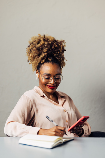 A smiling African-American entrepreneur with glasses writing something in her notebook while listening to some business podcast on her smartphone. She is sitting at the desk in her office. (copy space)