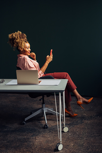 A side view of a smiling African-American entrepreneur chatting on her smartphone while sitting at the desk in her office. (copy space)