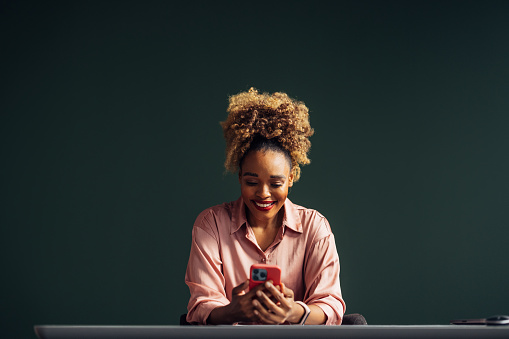 A smiling African-American entrepreneur watching something on her smartphone while sitting at the desk in her office. (copy space)