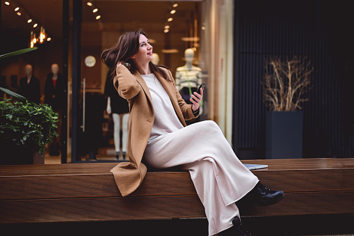 Fashionable mature woman in a brown trendy coat  sitting outdoors while having a break from a work in office. expressing positivity