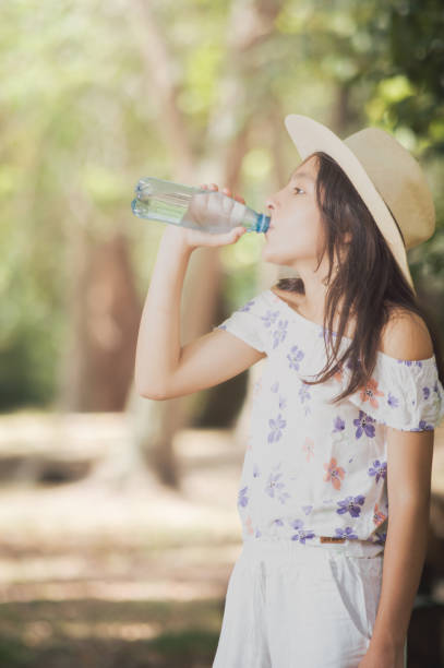 A girl drinks water while sitting on the beach by the lake during a summer day.