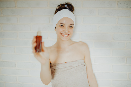 Portrait of a young Caucasian female wrapped in a towel holding a serum bottle and while standing against a white background.