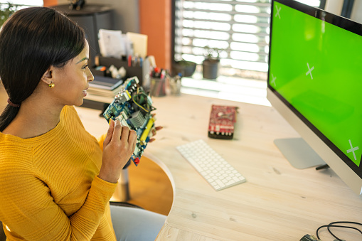 Hispanic female computer scientist sitting at her desk and showing motherboard to green screen during an online course.