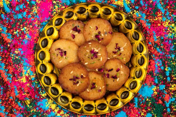 Sugary Pancakes Called Malpua, Pooa Or Pua Are Traditional Indian Dessert Mithai Drenched, Dunked, Soaked Or Dowsed In Cheeni Ki Chasni On Scattered Rainbow Color Powder Or Organic Gulal Background