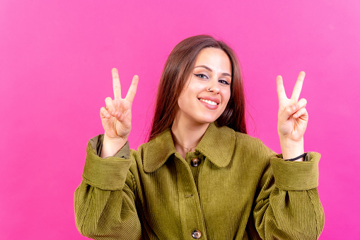 Young woman over pink background smiling and showing victory sign wearing green windbreaker