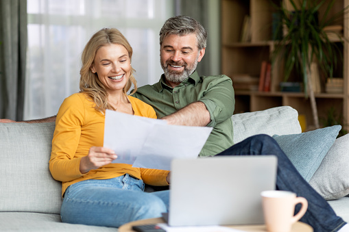 Family Insurance Concept. Happy Middle Aged Couple Reading Documents At Home, Smiling Mature Spouses Checking Financial Papers While Sitting Together On Couch In Living Room, Free Space