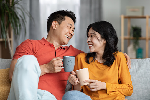Portrait of positive asian lovers happy man and woman in casual outfits sitting on sofa in cozy living room, drinking tea or coffee together, having conversation, looking at each other and laughing