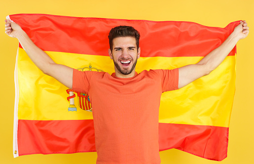 Happy caucasian man smiling and raising a spanish national flag in studio with yellow background