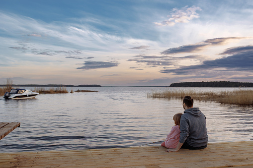 Father and daughter on the shore of the lake watching the sunset sitting on a wooden pier. Lifestyle, leisure, happiness, joy concept. Leisure.