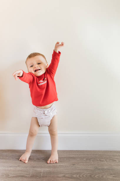 un lindo bebé de 14 meses lloriqueando y quejándose, de pie descalzo con los brazos extendidos y vistiendo un suéter rojo que dice "littlest valentine" con un pañal de tela de color nude mientras está en un piso de madera dura para el día de san vale - complaining whining no easel fotografías e imágenes de stock