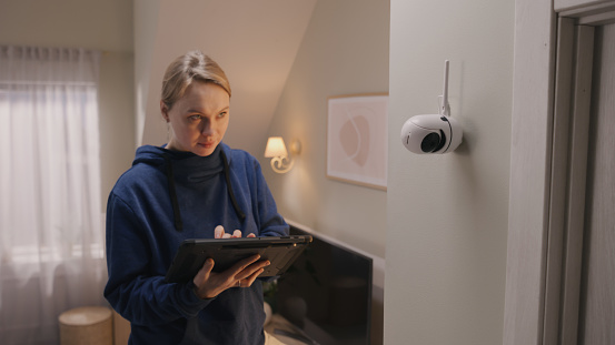 Caucasian woman installs security camera. Woman sets up angle of CCTV camera at home and rotates it with digital tablet computer and application. Concept of monitoring system, safety and privacy.