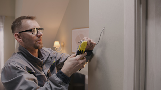 Man in uniform and glasses works with cable in drilled hole. Electrician removes insulation from wire with professional tool in the apartment. Installation of Internet or electricity work.