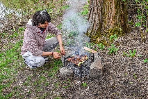 Young man frying meat on skewers on a campfire in the forest outdoors. Cooking during camping in spring. An adult guy is resting in a clearing on the shore of a pond