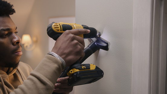 African American man in hoodie drills hole in concrete wall with hammer drill. Man does repairs in his apartment. Repair work, installation of the Internet or making hole for the picture.