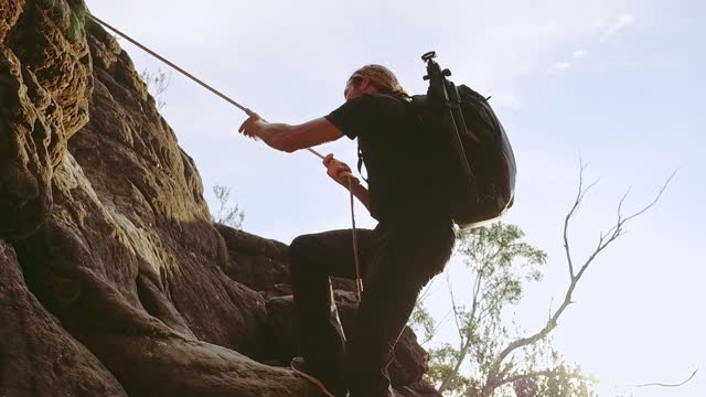 Young man using rope to climb up the edge of a mountain with backpack