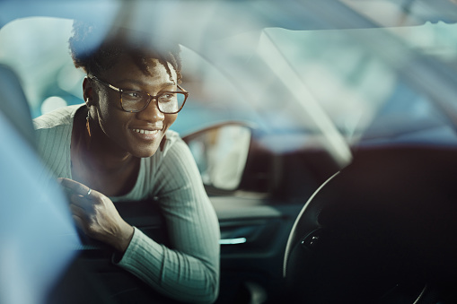 Happy African American woman looking at car interior while shopping in a showroom. The view is through glass.