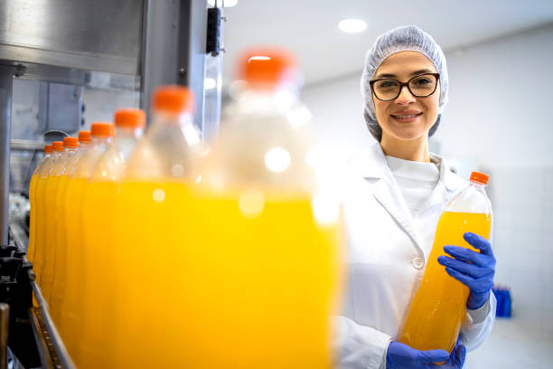 supervisora de fábrica de alimentos de pie en la planta embotelladora y sosteniendo una botella de jugo de naranja. - bottling plant industry drink food processing plant fotografías e imágenes de stock