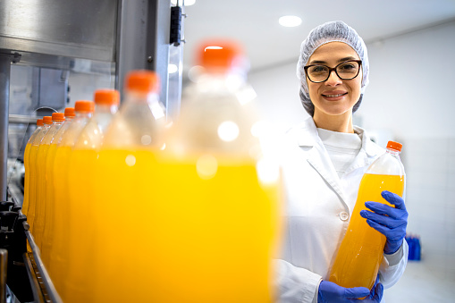 Food factory female supervisor standing in bottling plant and holding orange juice bottle.