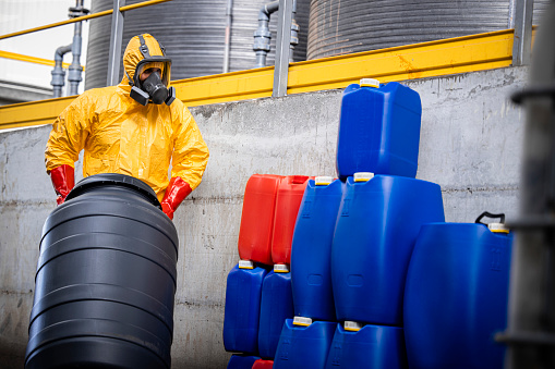 Chemical plant worker in protection suit moving barrel with chemicals in facility storage room.