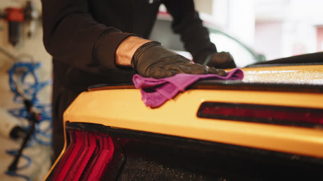 Cropped view of hand of male car wash worker holds the microfiber and polishes yellow sport luxury car under tail lights of vehicle. Car detailing and cleaning concept. Hand of male car wash worker.