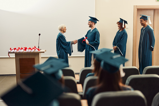 Happy senior professor shaking hands with her graduate students in a lecture hall. Copy space.