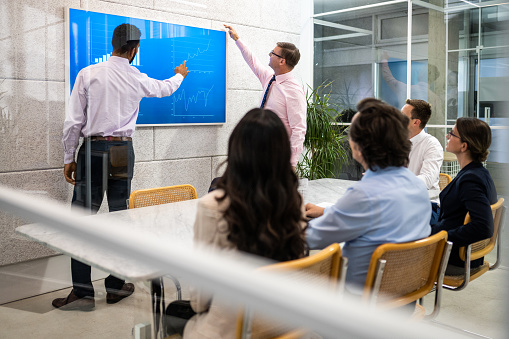 Corporate coworkers listening to financial statistics presentation in conference room. Business people discussing market research infographics on screen, back side view behind window