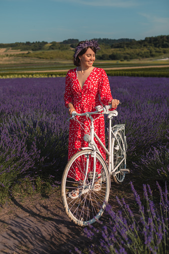 a young woman in a red dress stands with white bicycle in a lavender field