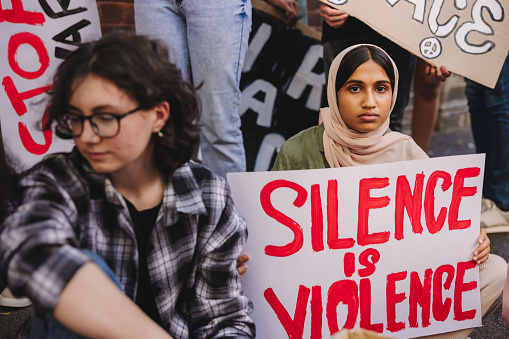 Young Muslim girl looking at the camera while holding an anti-violence poster. Diverse teenagers protesting against war and ethnic violence. Youth activists holding a peace and human rights demonstration.