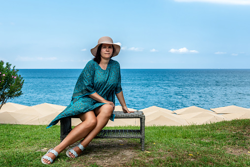 A young woman in a long dress and a wide-brimmed hat on a bench on the shores of the Mediterranean Sea