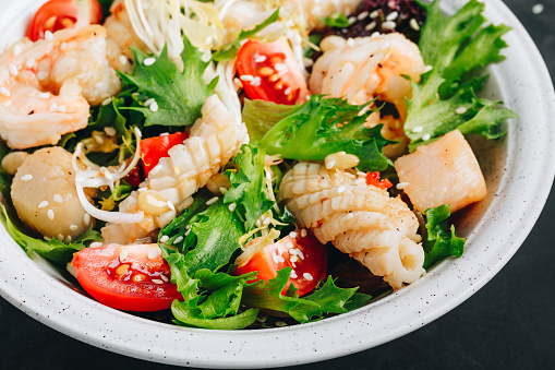 Seafood salad bowl. Green lettuce tomato salad with shrimps, scallop and squids on dark background