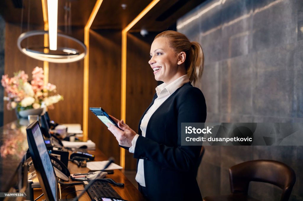 A happy receptionist is talking with hotel guest and making a reservation on a tablet. A friendly receptionist is talking to a client while making a reservation on a tablet at hotel reception. Hotel Stock Photo