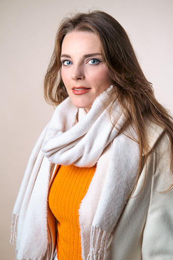 Studio shot of a woman in a white coat, white scarf and orange sweater. Winter fasihon.