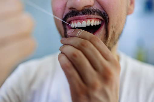 A close-up of a young Caucasian man flossing his teeth with a smile.