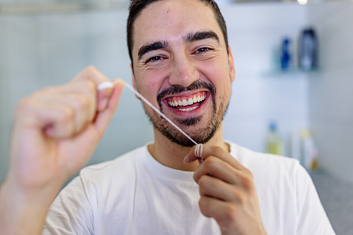 A young Caucasian man is looking at the camera with a big smile, while holding floss around his fingers.