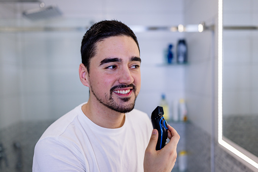 A young Caucasian man is checking himself out in the mirror, after shaving his beard with an electric shaver.
