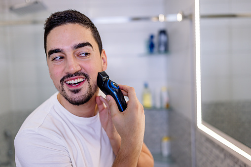 A young Caucasian man is using an electric shaver on his beard, while standing in a bathroom.