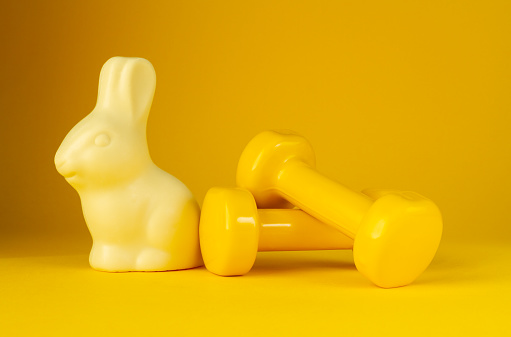 Chocolate Easter bunny with heavy dumbbells on yellow background. Healthy fitness lifestyle composition, gym workout and training concept. Cheat day temptation vs sticking to diet.